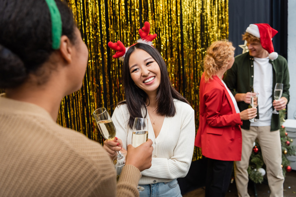 Lächelnde asiatische Geschäftsfrau hält Champagner in der Hand und spricht mit multiethnischen Kollegen während der Weihnachtsfeier im Büro  - Foto, Bild