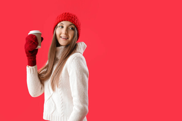 Młoda kobieta w ciepłym kapeluszu z filiżanką kawy na wynos na czerwonym tle - Zdjęcie, obraz