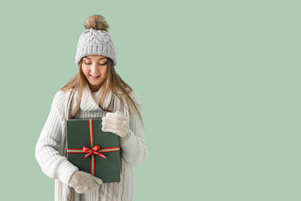 Young woman in warm hat with Christmas gift on green background - Foto, afbeelding