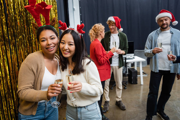 Positivo interracial businesswomen en Navidad diademas celebración champán cerca de oropel en oficina  - Foto, Imagen