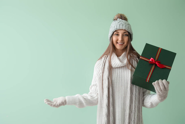 Młoda kobieta w ciepłym kapeluszu z prezentem świątecznym pokazującym coś na zielonym tle - Zdjęcie, obraz