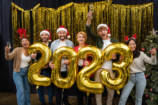 Pessoas de negócios inter-raciais alegres segurando champanhe e balões em forma de 2023 no escritório  - Foto, Imagem