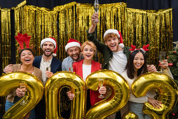 Positive multiethnic business people with champagne looking at camera near balloons in shape of 2023 sign in office  - 写真・画像