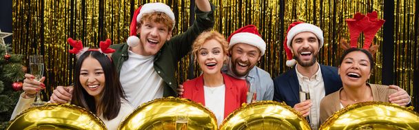 Cheerful multiethnic business people holding glasses of champagne near balloons during New Year party in office, banner  - Foto, Bild