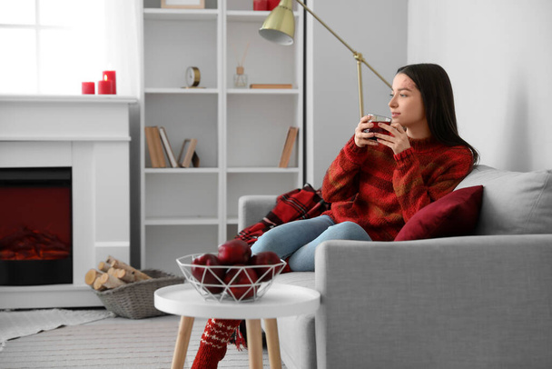 Молода жінка в теплому светрі зі скляною чашкою чаю сидить на дивані вдома
 - Фото, зображення