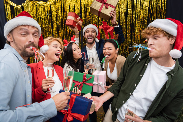 Smiling multiethnic business people in santa hats holding champagne and gifts near tinsel during party in office  - Photo, Image