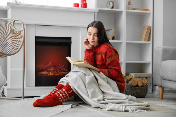 Młoda kobieta w ciepłym swetrze czyta książkę w pobliżu kominka w domu - Zdjęcie, obraz