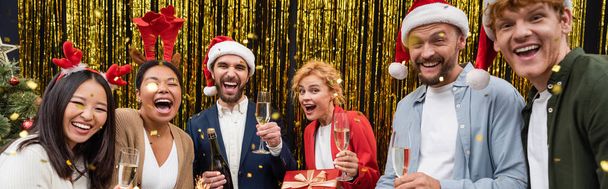 Positive interracial business people with champagne glasses standing under confetti during Christmas party in office, banner  - Photo, Image