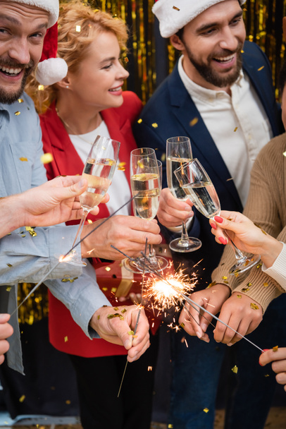 Cheerful multiethnic business people holding glasses of champagne and sparklers during Christmas party in office  - Fotó, kép