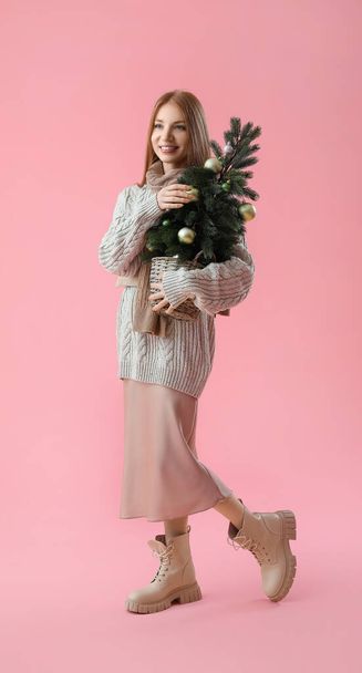 Young redhead woman in warm scarf with small Christmas tree on pink background - Photo, Image