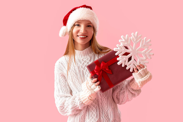 Młoda kobieta w kapeluszu Santa z prezentem świątecznym i płatkiem śniegu na różowym tle - Zdjęcie, obraz