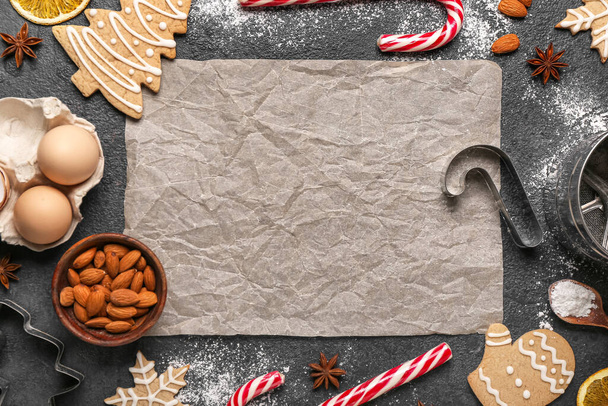 Composición con hoja de papel para hornear, galletas de Navidad e ingredientes sobre fondo oscuro - Foto, imagen