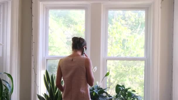 tylny widok materiał wideo młodej kobiety ze smartfonem przed oknem w domu - Materiał filmowy, wideo