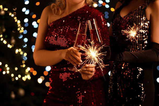 Γυναίκες σε κομψά φορέματα με φλεγόμενα sparklers και ποτήρια σαμπάνιας κατά θολή χριστουγεννιάτικα φώτα, closeup - Φωτογραφία, εικόνα