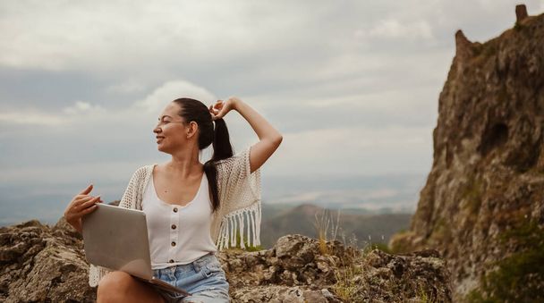 若い女性のフリーランスの旅行者はどこでもノートパソコンを使って山のピークビューを楽しんで屋外で働いている。幸せな女性ダウンシフターホールディングコンピュータで異常な素晴らしい景色の職場で夏 - 写真・画像