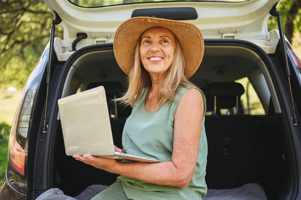 Senior happy woman working with laptop from the car trunk or boot in camper sunny summer park - digital nomad freedom lifestyle concept, travel freelancer and vacations of retired old age people. - Fotoğraf, Görsel