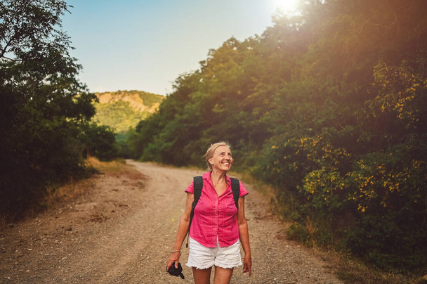 Podekscytowana szczęśliwa starsza kobieta turysta Backpacker spacery w letniej leśnej drodze na świeżym powietrzu o zachodzie słońca. Stara szczupła kobieta podróżująca z aparatem fotograficznym. Koncepcja aktywnego urlopu. Filtr ciepły - Zdjęcie, obraz