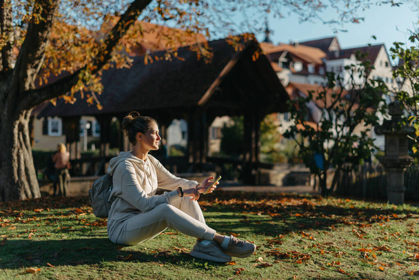Young fashionable teenage girl with smartphone in park in autumn sitting at smiling. Trendy young woman in fall in park texting. Retouched, vibrant colors. Beautiful blonde teenage girl wearing casual - Fotografie, Obrázek