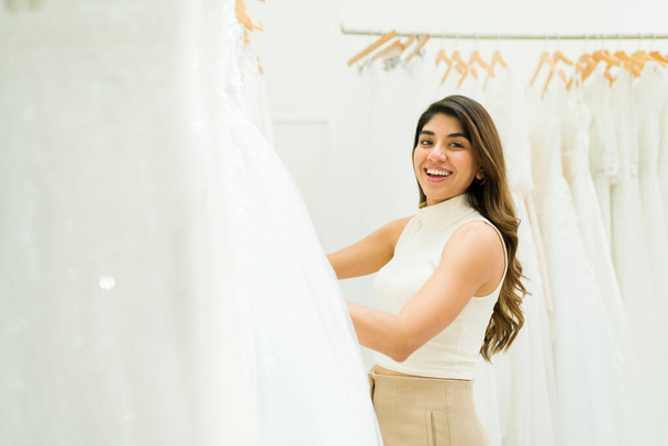 Beautiful hispanic woman looking excited while shopping for a wedding dress at the bridal shop - Photo, Image