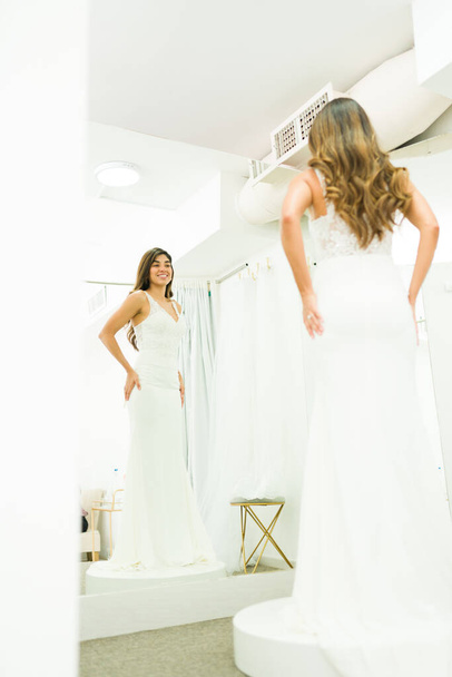 Mujer latina feliz vista desde atrás mirándose en el espejo en el vestidor mientras compra un vestido de novia - Foto, Imagen