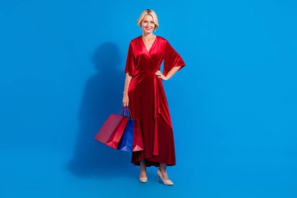 Full length photo of charming cute senior lady wear red shiny dress holding bargains isolated blue color background. - Foto, afbeelding