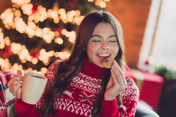 Photo of nice adorable sweet overjoyed girl with brunette hair wear red ornament jumper enjoy cookie hold tea cup in house indoors. - Fotó, kép
