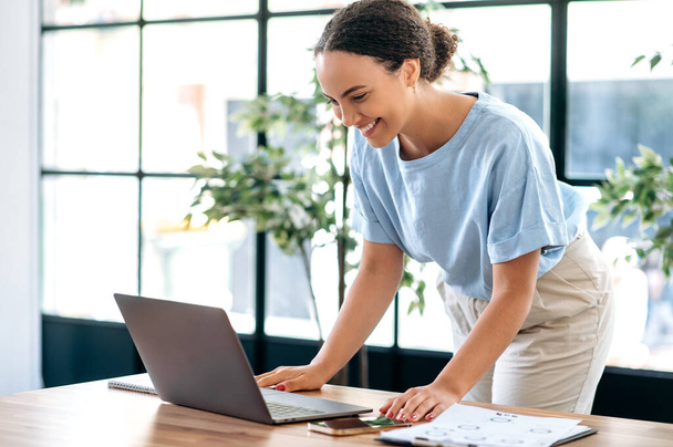Positive concentrated busy mixed race female employee, successful business lady, working with laptop, typing text, answering email, while standing in modern office near work desk, smiling - Foto, Bild