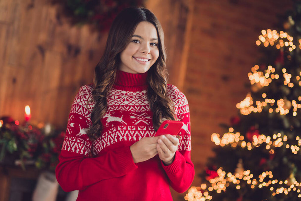 Portrait of cheerful satisfied good mood girl with wavy hairdo dressed red ornament sweater hold smartphone read post house indoors. - Foto, imagen