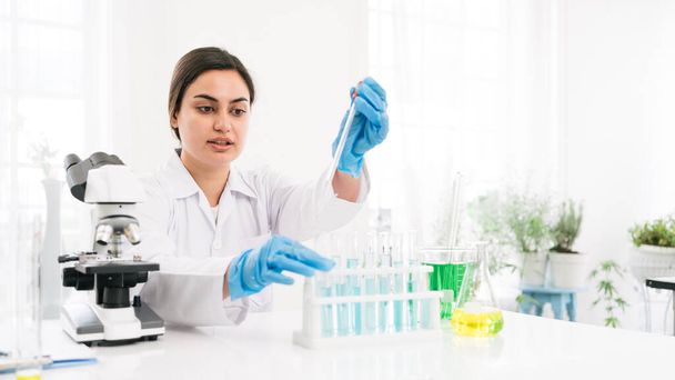 Een vrouwelijke onderzoeker uit het Midden-Oosten gebruikt een druppelaar om een stof in een reageerbuis te druppelen voor de analyse van vloeistoffen in het lab. Wetenschapper werkt met een druppelaar en een reageerbuis. - Foto, afbeelding