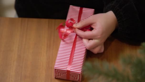 Женщина заворачивает подарок в раздетую бумагу с красной лентой. Руки упаковывают коробку в красно-белую рождественскую бумагу.  - Кадры, видео