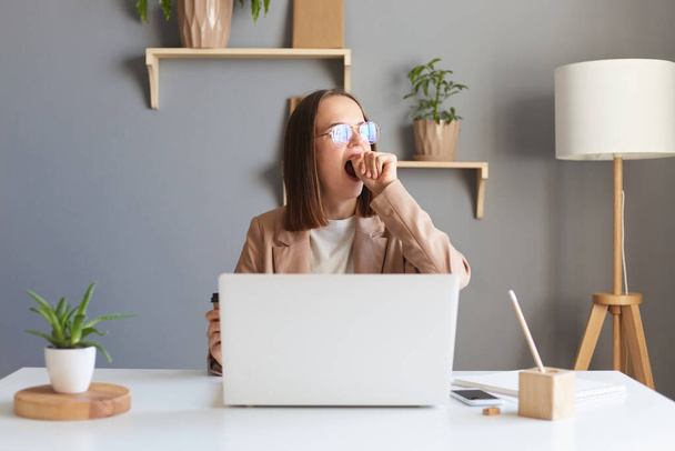 Attractive beautiful young Caucasian female feel tired and overworked, woman in glasses work remotely from home or sitting at table in office in front of laptop, yawning and covering mouth. - Foto, afbeelding