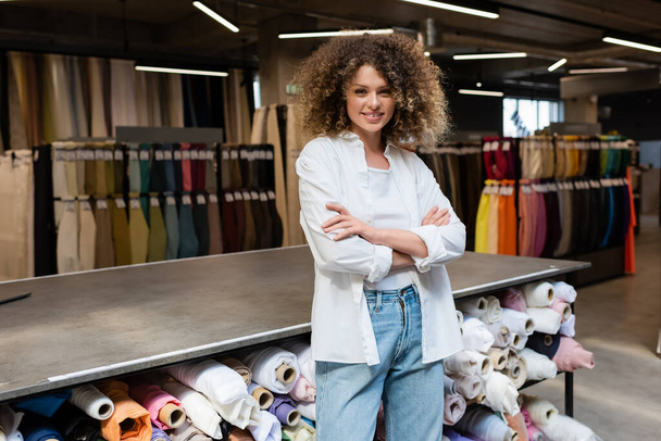 веселая и кудрявая продавщица, стоящая со скрещенными руками возле стойки с различными рулонами ткани в текстильном магазине  - Фото, изображение