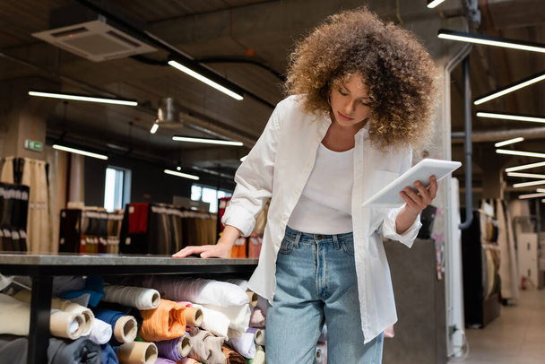 göndör értékesítő nő digitális táblagépet tart és szövettekercseket ellenőriz a textilboltban  - Fotó, kép