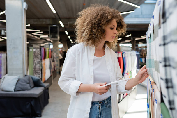 πλαϊνή άποψη του σγουρά πωλήτρια κρατώντας ψηφιακή ταμπλέτα, ενώ τον έλεγχο καρό ύφασμα στο κατάστημα κλωστοϋφαντουργίας  - Φωτογραφία, εικόνα