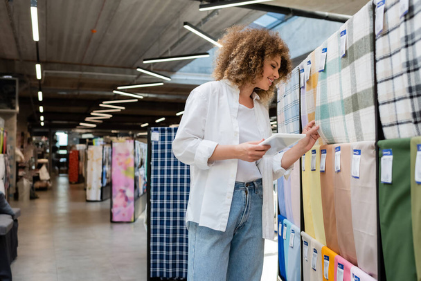 fröhliche Verkäuferin hält digitales Tablet in der Hand, während sie karierte Stoffe im Textilgeschäft überprüft  - Foto, Bild