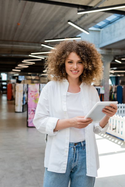 smiling saleswoman with curly hair holding digital tablet in textile shop  - Foto, Imagem
