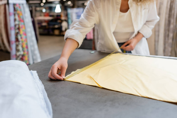 обрезанный вид продавщицы измеряющей желтый рулон льняной ткани в текстильном магазине  - Фото, изображение