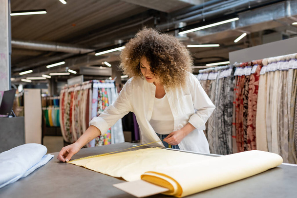 young and curly salesperson measuring yellow linen fabric roll in textile shop  - Foto, imagen