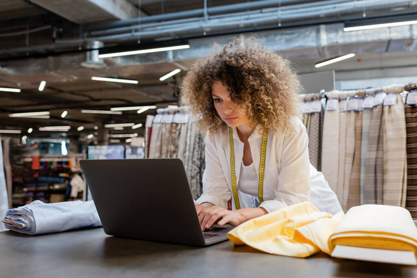 vendeuse bouclée avec ruban à mesurer à l'aide d'un ordinateur portable près de rouleaux de tissu dans un magasin de textiles  - Photo, image