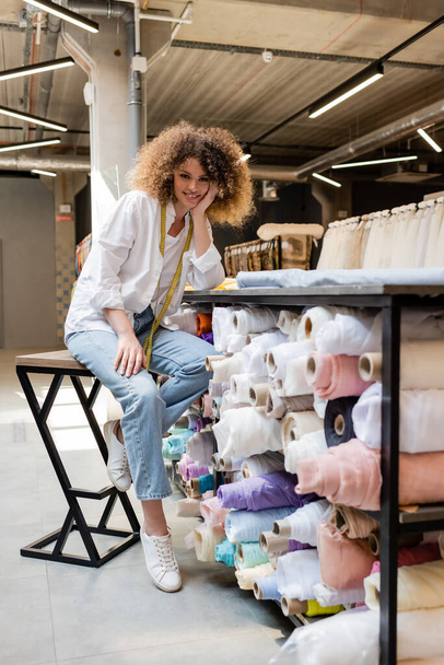vendedora alegre con cinta métrica sentada en la silla alta cerca del estante con rollos de tela en la tienda textil  - Foto, Imagen