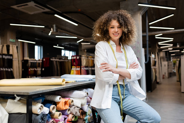 веселая продавщица с измерительной лентой, позирующая со скрещенными руками возле стойки с рулонами ткани в текстильном магазине  - Фото, изображение