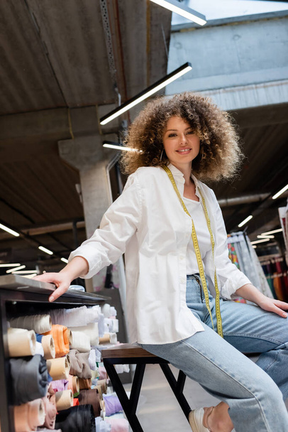 low angle view of joyful saleswoman with curly hair sitting near rack with fabric rolls in textile shop  - Photo, Image