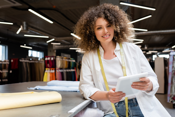 heureuse vendeuse tenant tablette numérique près des rouleaux de tissu sur le bureau dans le magasin de textile  - Photo, image