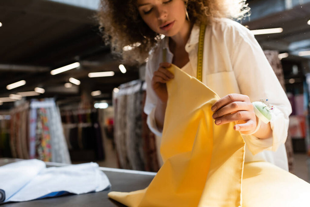 curly salesperson holding yellow fabric while working in textile shop  - Foto, Imagen