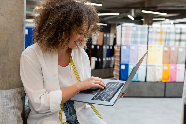 щасливий продавець з вимірювальною стрічкою за допомогою ноутбука під час роботи в текстильному магазині
  - Фото, зображення
