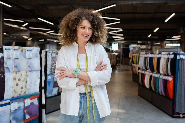 curly and cheerful saleswoman standing with crossed arms and looking away in textile shop - Photo, Image