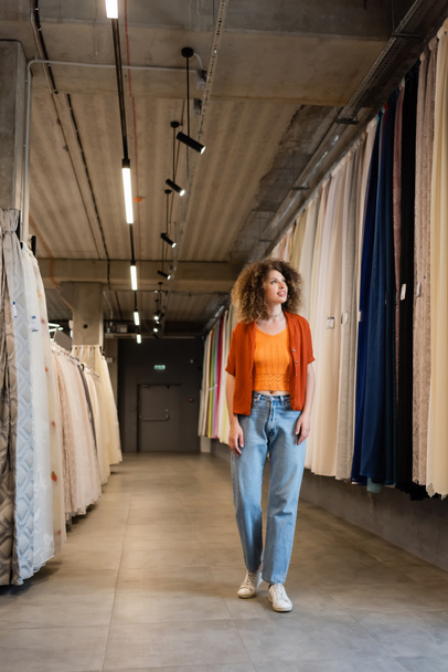 full length of curly woman in jeans walking along assortment of curtains and fabric in textile shop - Fotografie, Obrázek