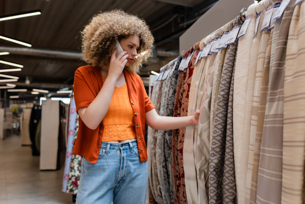 Кучерявий клієнт розмовляє по смартфону і вибирає тканину в текстильному магазині
  - Фото, зображення