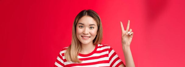 Close-up cheerful glad young attractive blond woman wear striped t-shirt smiling friendly make order, reservation for two show victory peace sign, stand red background, wanna be second. - Photo, Image