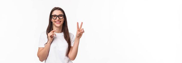 Entertainment, fun and holidays concept. Portrait of carefree cheerful young girl enjoying awesome party, celebrating show peace sign and wearing carnaval mask, standing white background. - Photo, Image
